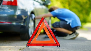 Red warning triangle sign on the road with a man checking his broken car in background