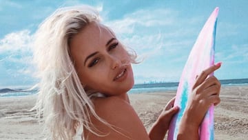 La surfista Ellie-Jean Coffey mirando a cámara y agarrándose a una tabla de surf rosa con una playa de fondo.