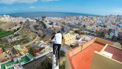 El piloto de MTB Danny MacAskill se pasea con su bici por una repisa de un tejado de Las Palmas (Gran Canaria, Islas Canarias, Espa&ntilde;a), con las famosas casas de colores del risco de San Juan a su derecha, la entrada a la ciudad delante y el mar al fondo. En diciembre del 2015. 
